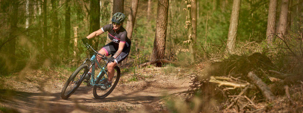 Vrouwelijke mountainbikester met roze 12GO Biking fietsshirt en lichtblauw MTB in een Nederlands bos op droge trail