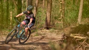 Vrouwelijke mountainbikester met roze 12GO Biking fietsshirt en lichtblauw MTB in een Nederlands bos op droge trail