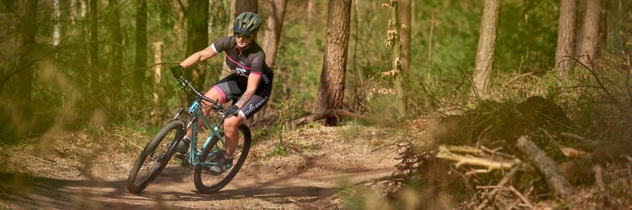 Vrouwelijke mountainbikester met roze 12GO Biking fietsshirt en lichtblauw MTB in een Nederlands bos op droge trail