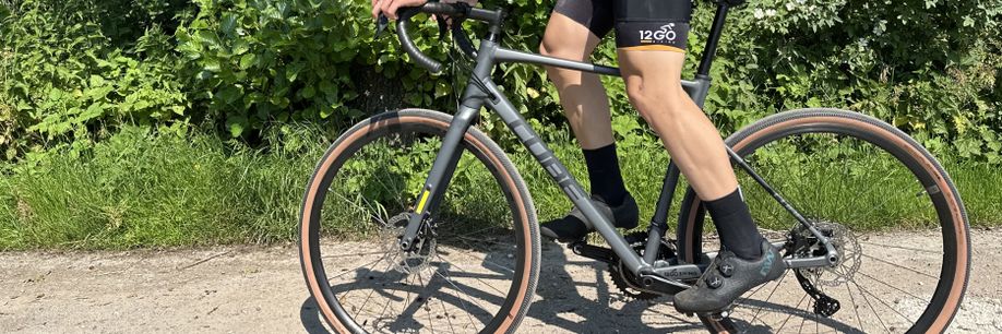 Fietser staat op de pedalen van een Cube Nuroad race gravelbike terwijl hij over een gravelpad fietst langs gras en struiken