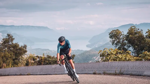 Man met blauw fietsshirt en zwarte racefiets rijdt richting camera de berg op met een fantastisch uitzicht met meer en bergen erachter