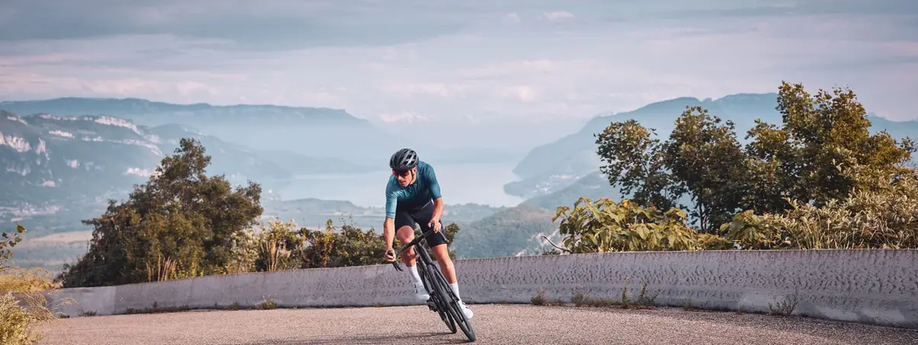 Man met blauw fietsshirt en zwarte racefiets rijdt richting camera de berg op met een fantastisch uitzicht met meer en bergen erachter