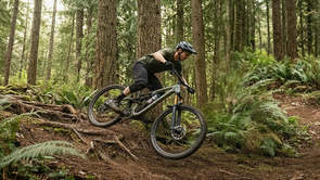 Moutainbiker met groene kleding op een groene mountainbike op een trail in een bos waarbij hij van de wortels afkomt en richting een bocht gaat