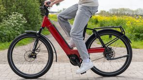 Persoon op een rode Riese & Müller Nevo e-bike die fietst over een klinkerweg met gravelpad erachter tussen struiken en planten ernaast