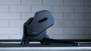 Tacx Flux 2 fietstrainer op een zwarte Tacx trainermat voor een betongetegelde muur