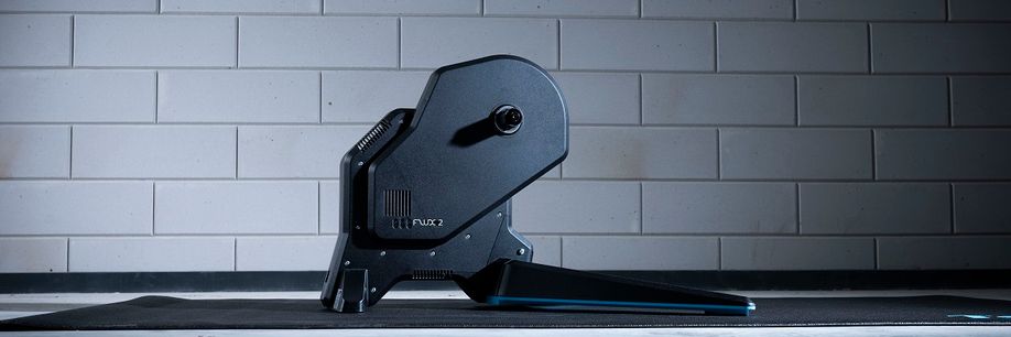 Tacx Flux 2 fietstrainer op een zwarte Tacx trainermat voor een betongetegelde muur