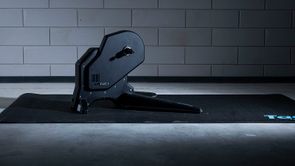 Tacx Flux S fietstrainer op zwarte Tacx trainermat met betongetegelde muur op de achtergrond