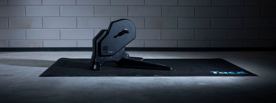 Tacx Flux S fietstrainer op zwarte Tacx trainermat met betongetegelde muur op de achtergrond