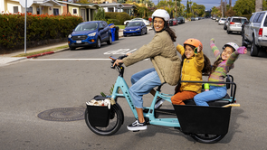 Vrouw met blauwe Trek Fetch longtail met twee kinderen achterop steekt kruispunt over met geparkeerde auto's in de zijweg