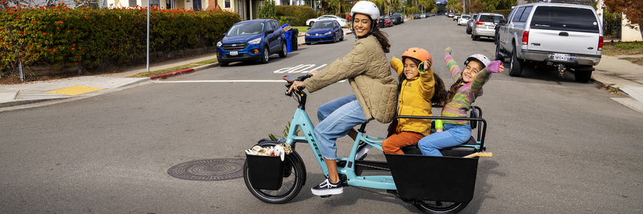 Vrouw met blauwe Trek Fetch longtail met twee kinderen achterop steekt kruispunt over met geparkeerde auto's in de zijweg