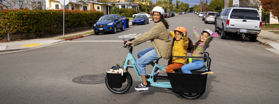 Vrouw met blauwe Trek Fetch longtail met twee kinderen achterop steekt kruispunt over met geparkeerde auto's in de zijweg