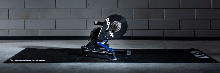 Wahoo Kickr Power V6 fietstrainer op een zwarte Wahoo trainermat met betongetegelde muur erachter