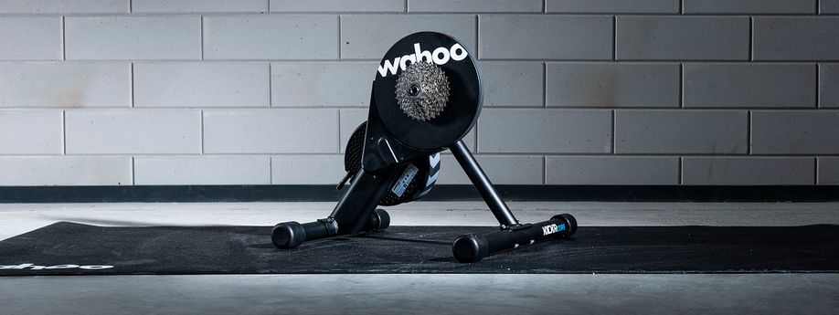 Wahoo Kickr Core fietstrainer op een zwarte Wahoo trainermat met betongetegelde muur als achtergrond