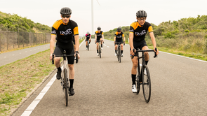 Groepje racefietsers met 12GO Biking fietskleding die verspreid naast en achter elkaar door de duinen rijden op een geasfalteerde autoweg