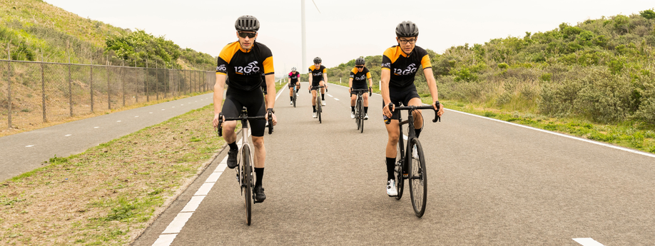Groepje racefietsers met 12GO Biking fietskleding die verspreid naast en achter elkaar door de duinen rijden op een geasfalteerde autoweg