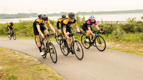 Groep racefietsers met 12GO Biking fietskleding die samen een heuveltje omhoog rijden met struiken erachter en een groot meer