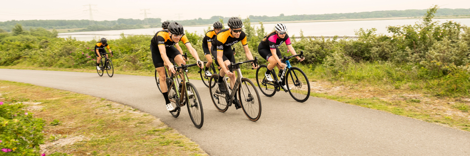 Groep racefietsers met 12GO Biking fietskleding die samen een heuveltje omhoog rijden met struiken erachter en een groot meer