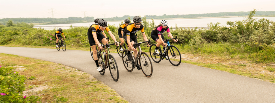 Groep racefietsers met 12GO Biking fietskleding die samen een heuveltje omhoog rijden met struiken erachter en een groot meer