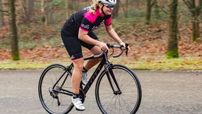 Vrouwelijke racefietser met 12GO Biking fietskleding fietst met zwarte Scott racefiets over asfaltweg langs de bossen