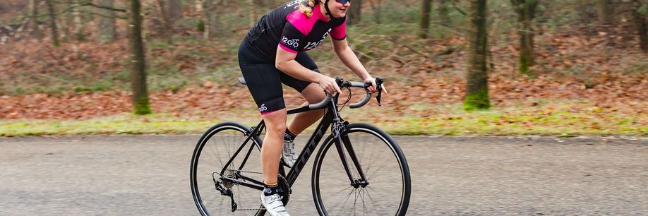 Vrouwelijke racefietser met 12GO Biking fietskleding fietst met zwarte Scott racefiets over asfaltweg langs de bossen