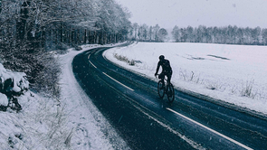 Fietser met lange fietskleding op racefiets over een donkere asfaltweg met sneeuw op een weiland rechts en witte sneeuwbomen links