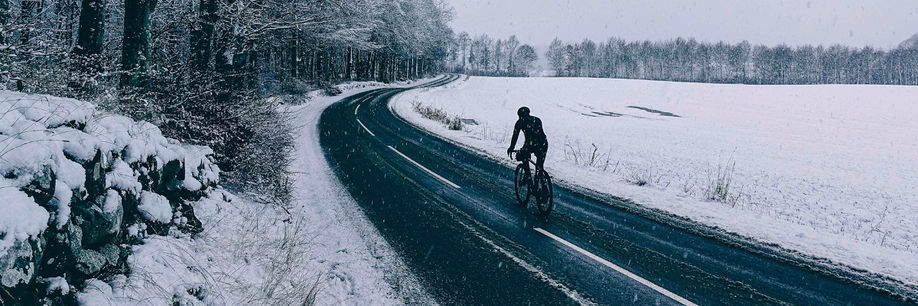 Fietser met lange fietskleding op racefiets over een donkere asfaltweg met sneeuw op een weiland rechts en witte sneeuwbomen links