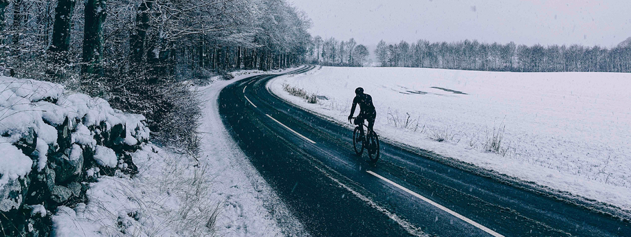 Fietser met lange fietskleding op racefiets over een donkere asfaltweg met sneeuw op een weiland rechts en witte sneeuwbomen links