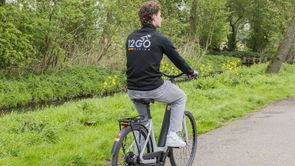 Man met 12GO Biking vest op grijszwarte e-bike en fiets over fietspad langs gras en klein slootje