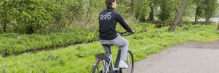 Man met 12GO Biking vest op grijszwarte e-bike en fiets over fietspad langs gras en klein slootje
