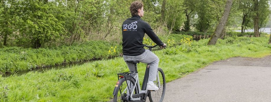 Man met 12GO Biking vest op grijszwarte e-bike en fiets over fietspad langs gras en klein slootje