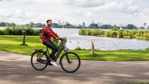 Man met rood lange mouwen shirt lacht op grijze e-bike terwijl hij op het asfalt van het Kralingse Bos fiets met water en de skyline erachter