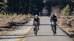 Twee fietsers met winterkleding op gravelbikes en 2 witte voorlichten over een asfaltweg tussen de heide