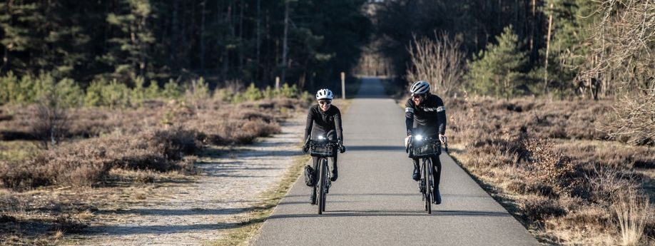 Twee fietsers met winterkleding op gravelbikes en 2 witte voorlichten over een asfaltweg tussen de heide