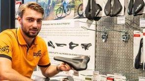 12GO Biking verkoper houdt fietszadel vast en kijkt de camera in voor een rek met veel fietszadels