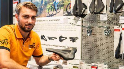 12GO Biking verkoper houdt fietszadel vast en kijkt de camera in voor een rek met veel fietszadels