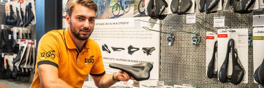 12GO Biking verkoper houdt fietszadel vast en kijkt de camera in voor een rek met veel fietszadels