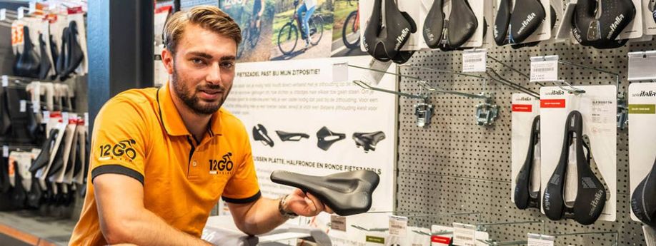 12GO Biking verkoper houdt fietszadel vast en kijkt de camera in voor een rek met veel fietszadels