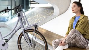 Vrouw op bankje binnen met paarse Cortina transportfiets met rekje voorop