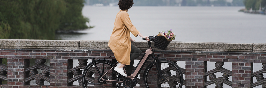 Vrouw met roze Kalkhoff fiets heeft bloemen in haar mandje voorop en staat op een stenen brug over de rivier uit te kijken