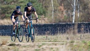 Twee fietsers met 12GO Biking fietskleding op gravelbikes over droog en vlak bospad met bomen en een stenen muur op de achtergrond