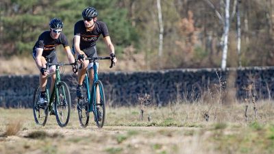 Twee fietsers met 12GO Biking fietskleding op gravelbikes over droog en vlak bospad met bomen en een stenen muur op de achtergrond