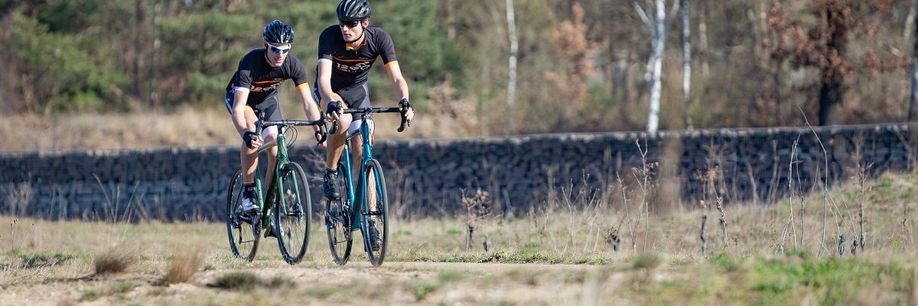 Twee fietsers met 12GO Biking fietskleding op gravelbikes over droog en vlak bospad met bomen en een stenen muur op de achtergrond