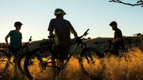 3 mountainbikers leunen op hun mountainbikes in het hoge gras terwijl de zon achter de bergen verdwijnt