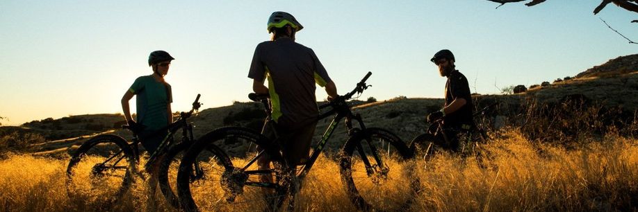 3 mountainbikers leunen op hun mountainbikes in het hoge gras terwijl de zon achter de bergen verdwijnt