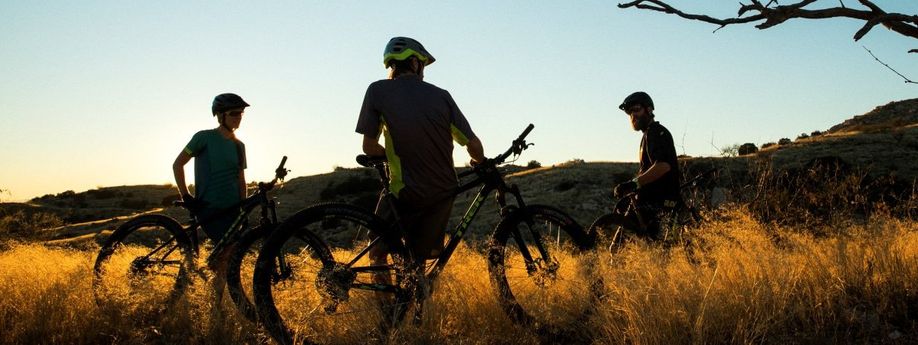 3 mountainbikers leunen op hun mountainbikes in het hoge gras terwijl de zon achter de bergen verdwijnt