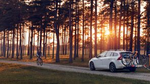 Man plaatst oranje mountainbike op fietsendrager achterop zijn witte auto met veel bomen en een ondergaande zon erachter