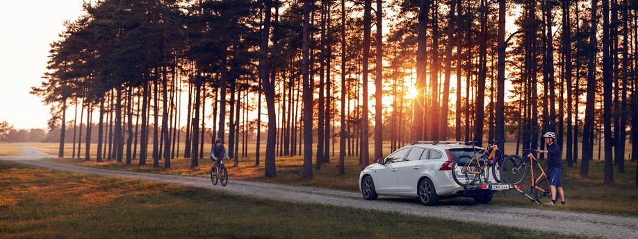 Man plaatst oranje mountainbike op fietsendrager achterop zijn witte auto met veel bomen en een ondergaande zon erachter