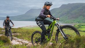 Vrouw met mountainbike en Fox broek rijd voor man met mountainbike een trail omhoog de bergen in met een meer en hoge bergen op de achtergrond