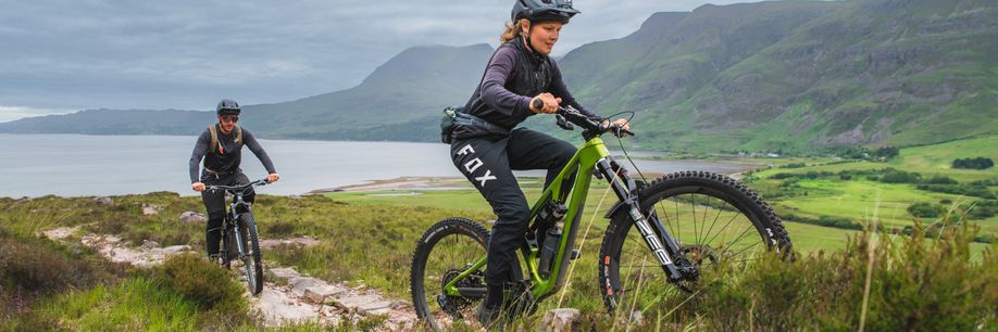 Vrouw met mountainbike en Fox broek rijd voor man met mountainbike een trail omhoog de bergen in met een meer en hoge bergen op de achtergrond