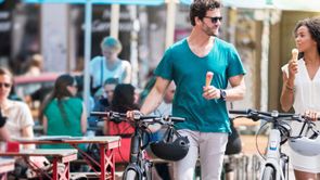 Man en vrouw lopen met fietsen in hand langs het terras terwijl ze in de andere hand beiden een ijsje hebben en met elkaar praten